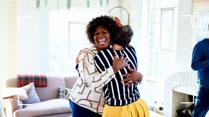 Woman laughing and smiling standing in living room hugging another woman with back to the camera, representing friendships and how to be happier